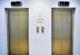 Two closed elevator doors are positioned side by side, set against a light-colored wall. Above each elevator is a digital display showing the letter 'L'. Between the elevators are control panels with buttons, and there are two small notes attached to the wall, each with visible text.