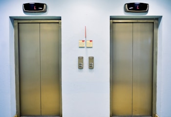 Two closed elevator doors are positioned side by side, set against a light-colored wall. Above each elevator is a digital display showing the letter 'L'. Between the elevators are control panels with buttons, and there are two small notes attached to the wall, each with visible text.