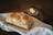 A loaf of freshly baked bread rests on a white cloth placed on a wooden surface. Beside it, there is a small glass dish with a piece of butter and a knife lying next to it.
