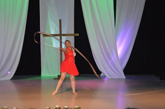 A performer in a red dress dances energetically with a ribbon on a stage. In the background, a large wooden cross is positioned against draped white fabric with colored lighting creating a vibrant atmosphere.