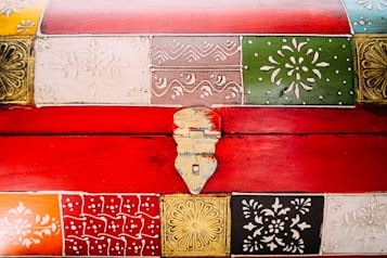 A vibrant, ornately decorated chest featuring panels with intricate patterns. The designs include floral and geometric motifs, each painted in different colors like red, green, brown, white, and black. The central metallic latch adds an antique touch.
