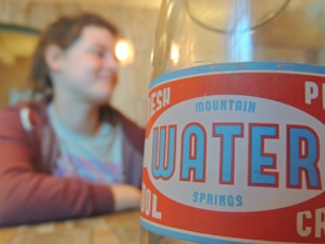 A close-up of a vintage-style bottle label for 'Mountain Springs Water' is in focus, while a person in casual attire is blurred in the background.