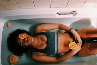 A person is lying in a bathtub filled with water, with slices of oranges placed on their body and around them. The person is wearing a dark sports bra and appears relaxed with eyes closed. The setting is minimalistic with white tiles surrounding the tub.