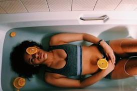 A person is lying in a bathtub filled with water, with slices of oranges placed on their body and around them. The person is wearing a dark sports bra and appears relaxed with eyes closed. The setting is minimalistic with white tiles surrounding the tub.