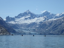 A serene landscape featuring towering snow-capped mountains with rugged peaks surrounded by glaciers. The foreground shows a calm body of water with several kayakers paddling towards the mountains, creating a sense of adventure and exploration. The scene is set under a clear blue sky.