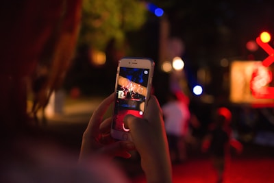 A person is holding a smartphone, capturing a scene in a dimly lit outdoor setting. The phone screen displays a blurred image of lights, possibly from a performance or installation. Surrounding the person, blurred figures and bright lights contribute to a lively nighttime atmosphere.
