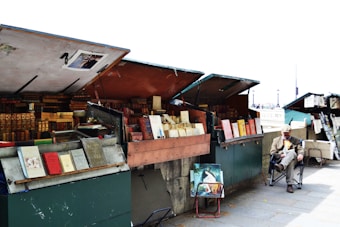 An outdoor book market with several open stalls displaying various books, some with ornate spines. A man sits on a chair next to one of the stalls, wearing a hat and a coat. A few paintings are leaning against the stalls.