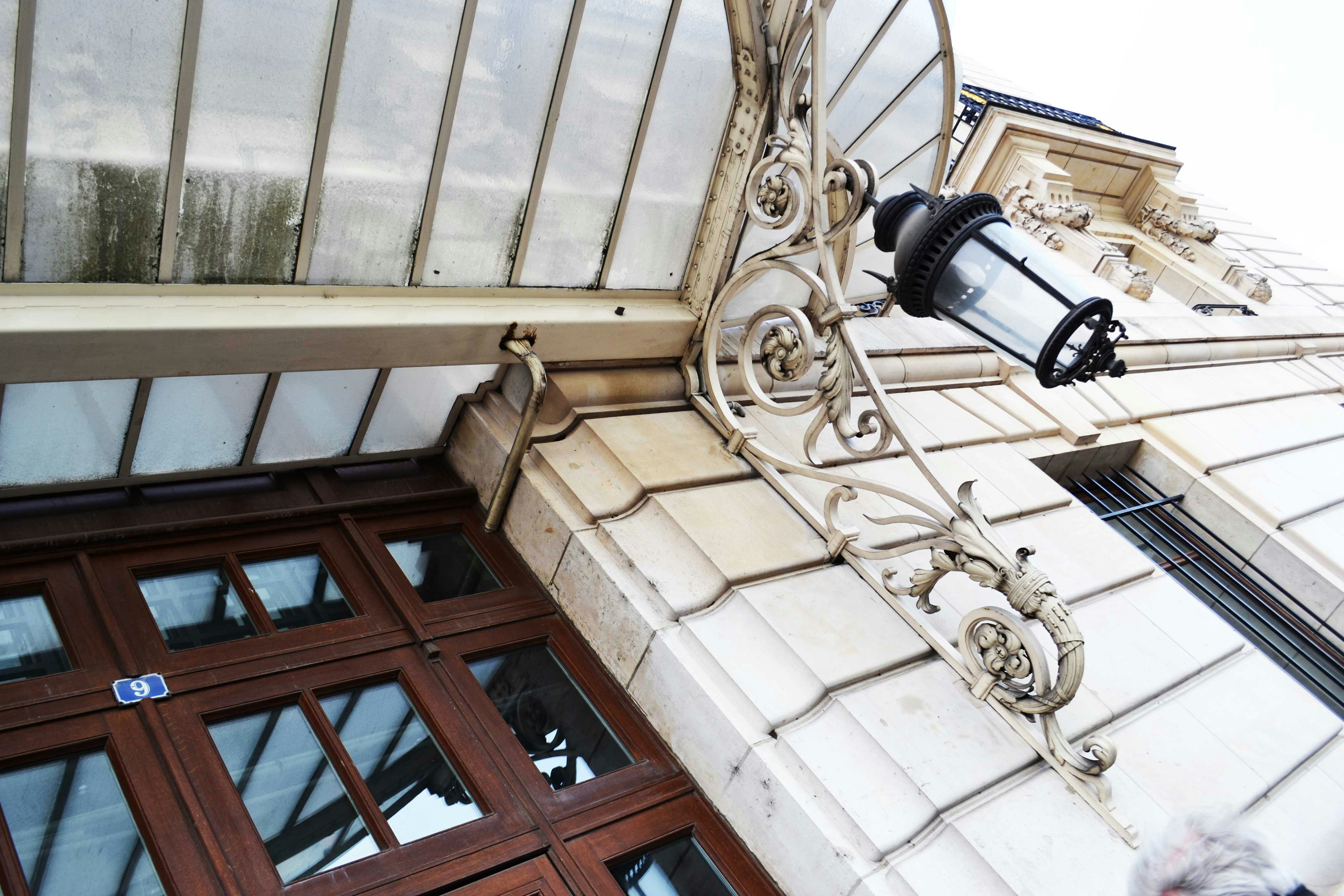 An ornate glass canopy with a wrought iron bracket and a vintage lantern attached to a stone building facade. Wooden framed windows with reflective glass panes are visible, alongside architectural details like molding work and stone patterns.