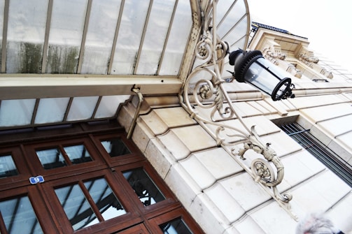 An ornate glass canopy with a wrought iron bracket and a vintage lantern attached to a stone building facade. Wooden framed windows with reflective glass panes are visible, alongside architectural details like molding work and stone patterns.