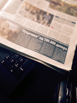 A newspaper page with an article featuring a photograph and text in a foreign language. The page is placed atop a black computer keyboard, with headphones partially visible on the side.