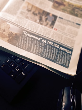 A newspaper page with an article featuring a photograph and text in a foreign language. The page is placed atop a black computer keyboard, with headphones partially visible on the side.