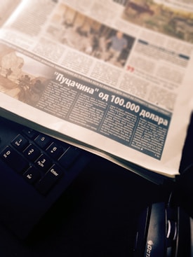 A newspaper page with an article featuring a photograph and text in a foreign language. The page is placed atop a black computer keyboard, with headphones partially visible on the side.