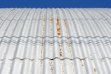 A close-up view of a corrugated metal roof with visible rust and weathering. The roof is casting wavy shadows due to its ridged surface, and the background is a clear blue sky.