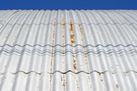 A close-up view of a corrugated metal roof with visible rust and weathering. The roof is casting wavy shadows due to its ridged surface, and the background is a clear blue sky.