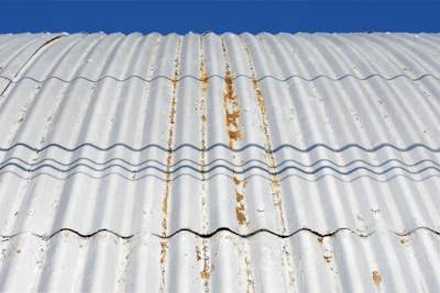 A close-up view of a corrugated metal roof with visible rust and weathering. The roof is casting wavy shadows due to its ridged surface, and the background is a clear blue sky.