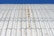 A close-up view of a corrugated metal roof with visible rust and weathering. The roof is casting wavy shadows due to its ridged surface, and the background is a clear blue sky.