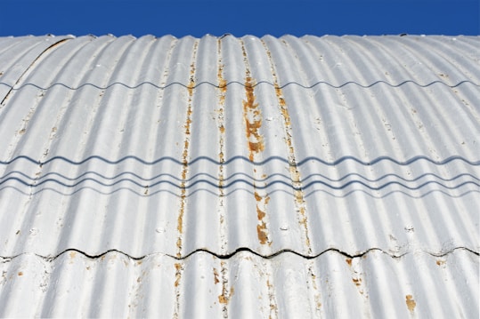 A close-up view of a corrugated metal roof with visible rust and weathering. The roof is casting wavy shadows due to its ridged surface, and the background is a clear blue sky.