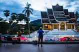 A traditional temple with intricate architecture and gold details stands prominently in the center. Surrounding it are tall palm trees and a mountainous backdrop, under a dramatic evening sky. People are seen taking photographs and viewing the scene from a distance, suggesting tourism or leisurely activity.