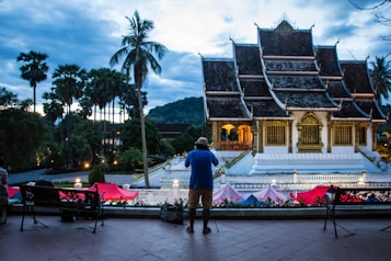 A traditional temple with intricate architecture and gold details stands prominently in the center. Surrounding it are tall palm trees and a mountainous backdrop, under a dramatic evening sky. People are seen taking photographs and viewing the scene from a distance, suggesting tourism or leisurely activity.