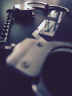 A pair of metallic handcuffs is placed on a textured dark surface. The handcuffs have a reflective sheen, and the image captures a close-up perspective with a shallow depth of field.