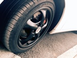 A close-up view of a car tire and rim, featuring a sleek black finish on the rim with visible tread patterns on the rubber tire. The background includes a portion of a white car body and an asphalt surface, adding contrast and texture to the scene.