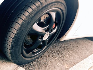 A close-up view of a car tire and rim, featuring a sleek black finish on the rim with visible tread patterns on the rubber tire. The background includes a portion of a white car body and an asphalt surface, adding contrast and texture to the scene.