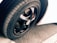 A close-up view of a car tire and rim, featuring a sleek black finish on the rim with visible tread patterns on the rubber tire. The background includes a portion of a white car body and an asphalt surface, adding contrast and texture to the scene.