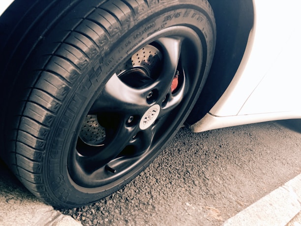 A close-up view of a car tire and rim, featuring a sleek black finish on the rim with visible tread patterns on the rubber tire. The background includes a portion of a white car body and an asphalt surface, adding contrast and texture to the scene.