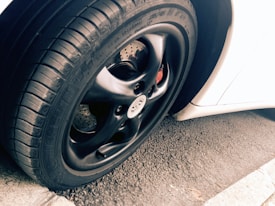 A close-up view of a car tire and rim, featuring a sleek black finish on the rim with visible tread patterns on the rubber tire. The background includes a portion of a white car body and an asphalt surface, adding contrast and texture to the scene.