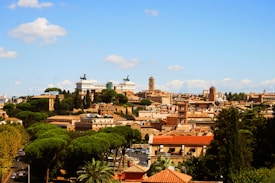 A panoramic view of a historic city with a mix of ancient and classical architecture. The skyline includes prominent buildings and monuments surrounded by lush greenery, suggesting a blend of urban development and natural landscape. The background features a clear blue sky with a few scattered clouds.