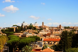 A panoramic view of a historic city with a mix of ancient and classical architecture. The skyline includes prominent buildings and monuments surrounded by lush greenery, suggesting a blend of urban development and natural landscape. The background features a clear blue sky with a few scattered clouds.