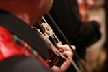 A close-up view of a person playing a violin. The focus is on the hand holding the instrument and the bow in motion, while a red plaid pattern is visible on part of the clothing. The background is slightly out of focus, adding a sense of depth to the image.
