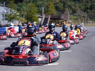 A group of people wearing helmets are driving go-karts lined up on a track, with the focus on the front kart. The setting appears to be an outdoor karting facility surrounded by greenery.