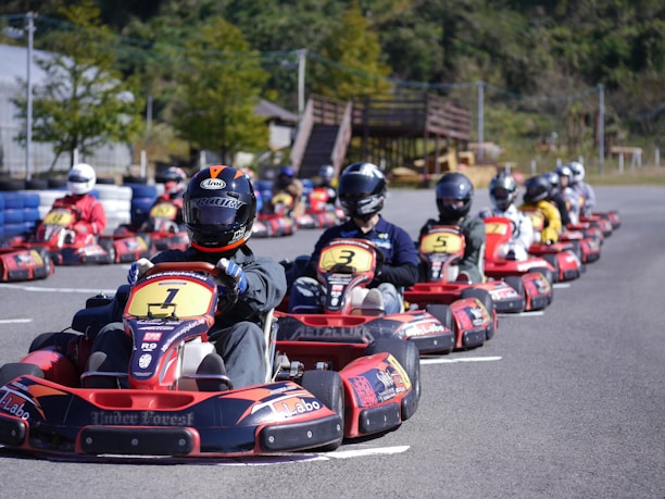 A group of people wearing helmets are driving go-karts lined up on a track, with the focus on the front kart. The setting appears to be an outdoor karting facility surrounded by greenery.