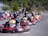A group of people wearing helmets are driving go-karts lined up on a track, with the focus on the front kart. The setting appears to be an outdoor karting facility surrounded by greenery.