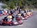A group of people wearing helmets are driving go-karts lined up on a track, with the focus on the front kart. The setting appears to be an outdoor karting facility surrounded by greenery.