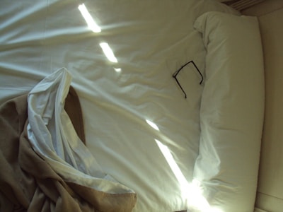 A bed with a neatly arranged white pillow and sheets is illuminated by natural light streaming through horizontal openings. A pair of glasses is placed on the pillow, and a brown blanket is casually draped at the edge.