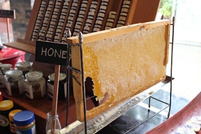 A large honeycomb displayed on a stand with the word 'honey' on a small chalkboard sign. Surrounding the honeycomb are various jars, possibly containing honey or spreads, and a collection of arranged small jars in the background.