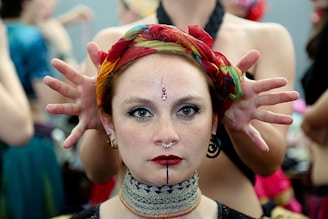 A woman with striking makeup and a decorative headpiece stares directly at the camera. Her hair is wrapped in a vibrant scarf with red, green, and yellow tones. Behind her, hands are posed as if framing her head. She wears bold makeup with red lipstick and facial jewelry, including a septum ring and an intricate bindu on her forehead.