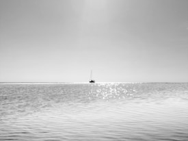 A minimalist black and white photograph featuring a lone sailboat on a vast expanse of water under a clear sky. The sunlight creates shimmering reflections on the water, adding a serene and contemplative mood.