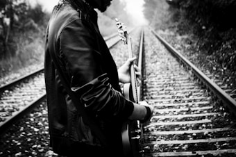 A person wearing a leather jacket is standing on a set of railway tracks holding a guitar. The scene is captured in black and white, creating a moody and contemplative atmosphere. The railway tracks lead into the distance, flanked by trees and foliage. The person is facing away, adding a sense of mystery.