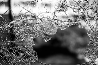 A piece of glass is shattered and has a large hole in the center. Jagged shards and fragments are scattered around the edges, with light reflecting off the uneven surfaces. The background is blurred, providing a contrast to the sharpness of the broken glass.
