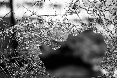 A piece of glass is shattered and has a large hole in the center. Jagged shards and fragments are scattered around the edges, with light reflecting off the uneven surfaces. The background is blurred, providing a contrast to the sharpness of the broken glass.