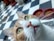 A close-up of a cat with green eyes and white and orange fur. The cat is looking upwards, and its whiskers are prominently visible. In the background, a black and white checkered floor is visible along with a red pet food bowl containing kibble.