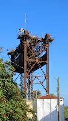 A tall industrial metal structure with a lattice design stands prominently against a clear blue sky. The rusty brown color of the metal contrasts with the greenery of a nearby tree, which is dotted with small red berries. A white structure with a red roof is located at the base of the metal tower, and power lines cross the image.