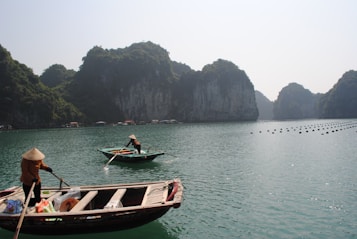 Two small boats navigated by people wearing traditional conical hats are set against a backdrop of towering limestone islands. The water is calm and dotted with buoys, suggesting a serene and tranquil environment. The landscape is lush with greenery while a slight mist floats in the background.
