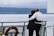 An older couple standing on a ferry deck, embracing while looking out over a body of water with a scenic view. The sky is cloudy, and the distant shoreline features a line of trees. A bright orange life ring is visible attached to the railing.