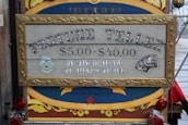 A vintage-style sign with ornate framing advertises a fortune teller service. The pricing ranges from $5.00 to $40.00 with the tagline 'Pay what you are able, the reading is the same.' The sign features mystical imagery, including an eye symbol and tarot cards. It's set against a colorful background with decorative elements.