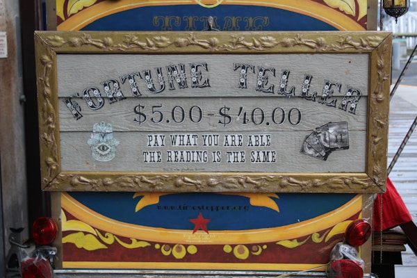 A vintage-style sign with ornate framing advertises a fortune teller service. The pricing ranges from $5.00 to $40.00 with the tagline 'Pay what you are able, the reading is the same.' The sign features mystical imagery, including an eye symbol and tarot cards. It's set against a colorful background with decorative elements.