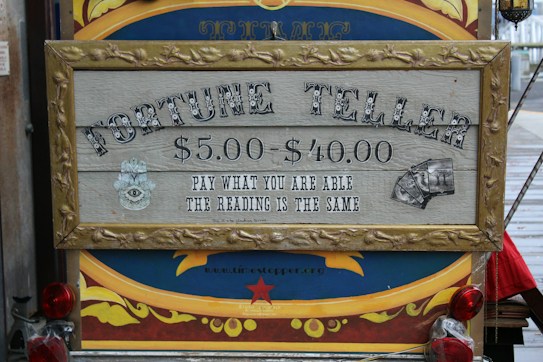 A vintage-style sign with ornate framing advertises a fortune teller service. The pricing ranges from $5.00 to $40.00 with the tagline 'Pay what you are able, the reading is the same.' The sign features mystical imagery, including an eye symbol and tarot cards. It's set against a colorful background with decorative elements.